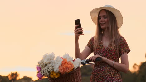 Beautiful-blonde-in-a-hat-with-a-bike-looking-at-the-mobile-phone-screen-and-a-basket-on-the-handlebar-with-flowers